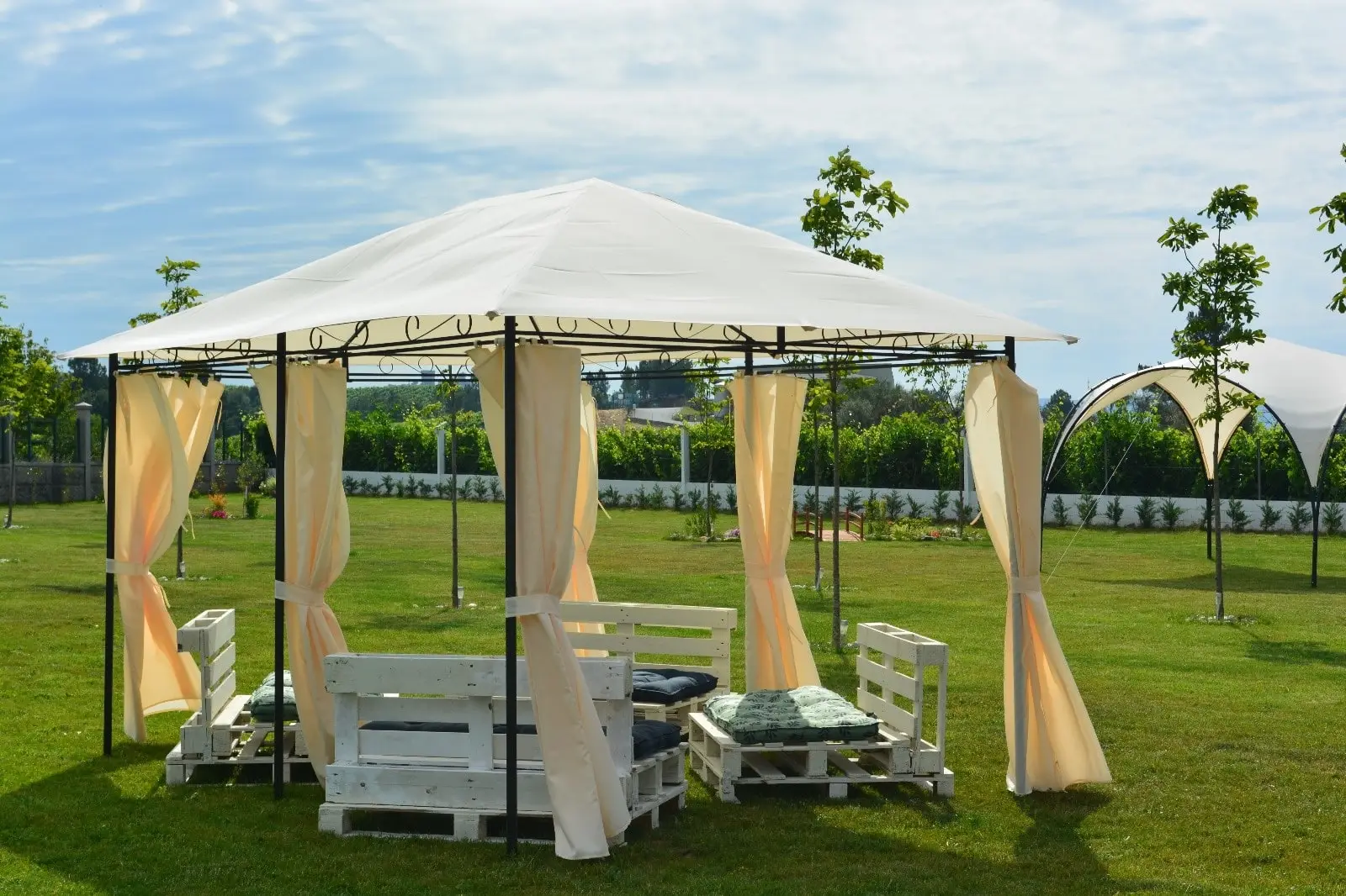 La glorieta del jardín, con cortinas crema recogidas, un sofá de palés blancos con cojines y el viñedo detrás