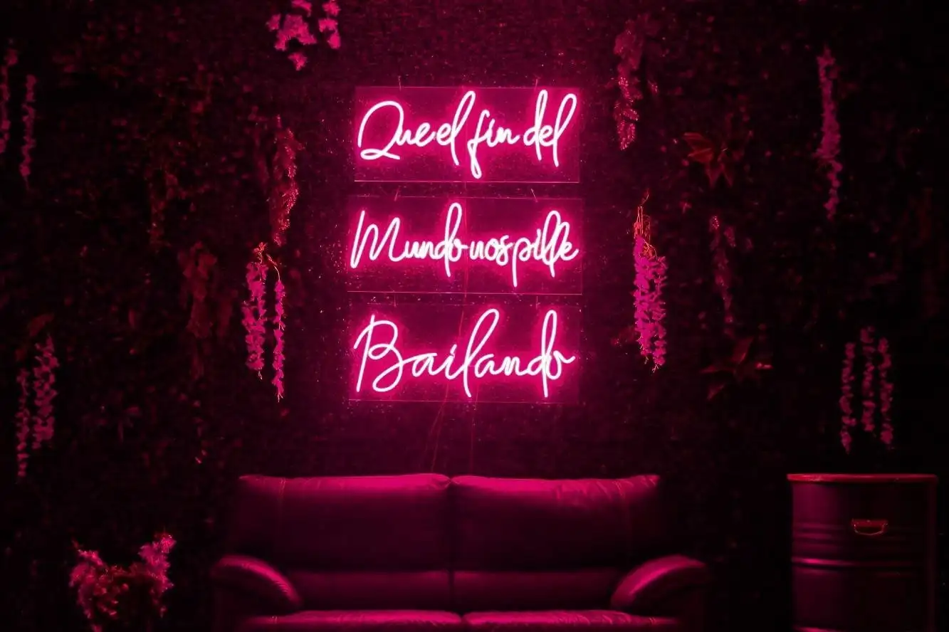 Un neón rosa con la frase «Que el fin del mundo nos pille bailando», sobre un muro de follaje, con un sofá rojo debajo
