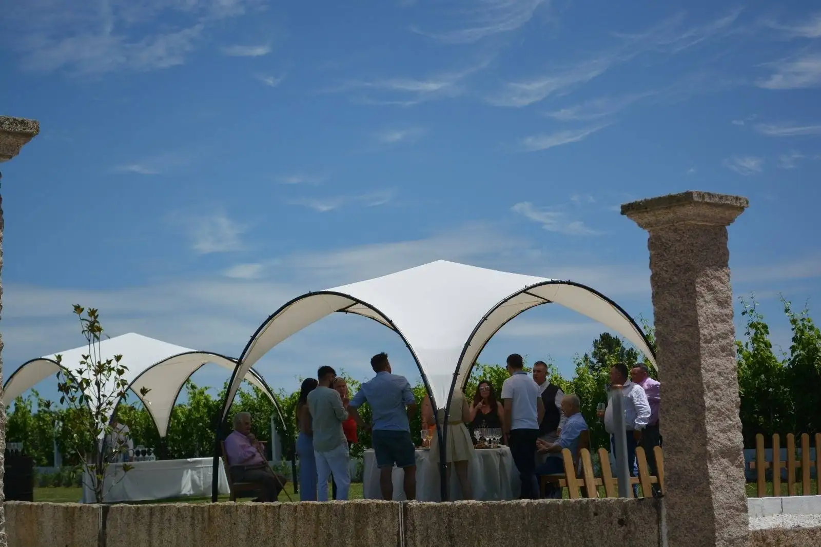 Un toldo blanco entre dos pilares de granito, con un muro de piedra y, debajo, invitados charlando