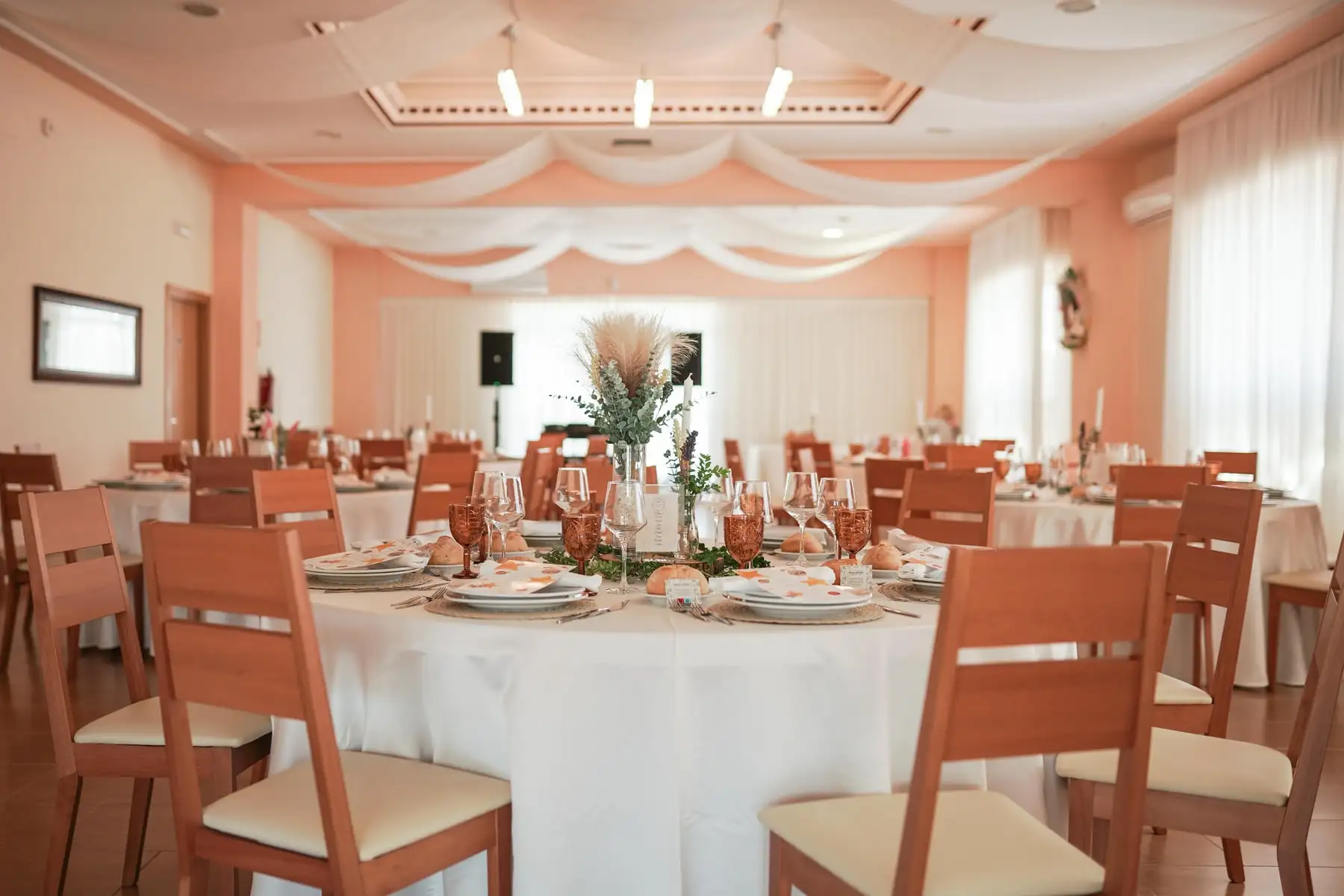 Un comedor con mesas redondas de mantel blanco, sillas de madera y telas blancas en el techo, con la luz de los ventanales