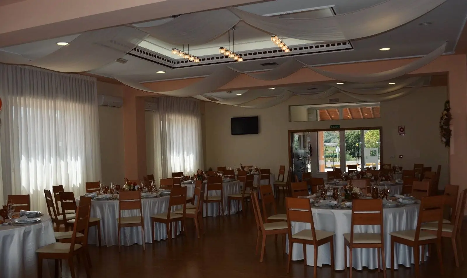 El comedor con mesas redondas de mantel blanco, suelo de madera y cortinas blancas en los ventanales