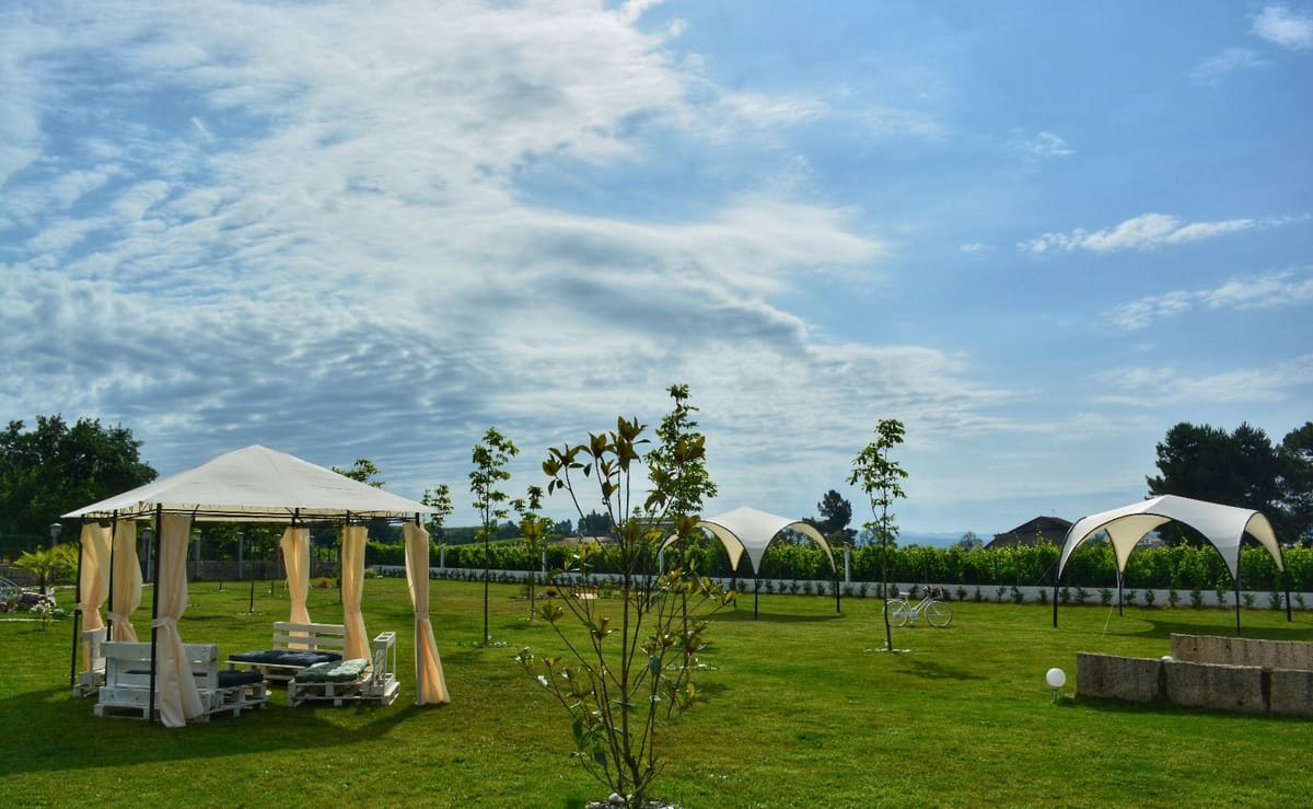El jardín de Finca Dona Branca: una glorieta de cortinas crema, dos toldos blancos, árboles jóvenes y el viñedo al fondo, bajo un cielo de nubes